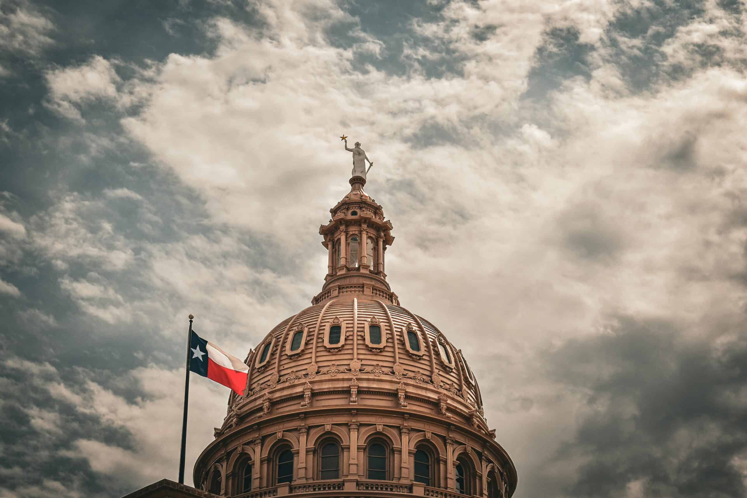 The dome of the Texas State Capitol with a Texas flag under a cloudy sky.