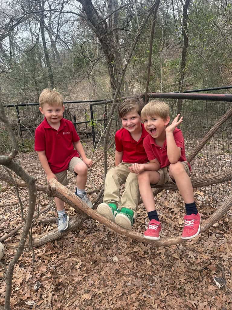 Three Selwyn School students in red uniform polos sitting on a fallen branch in the woods on the Argyle campus