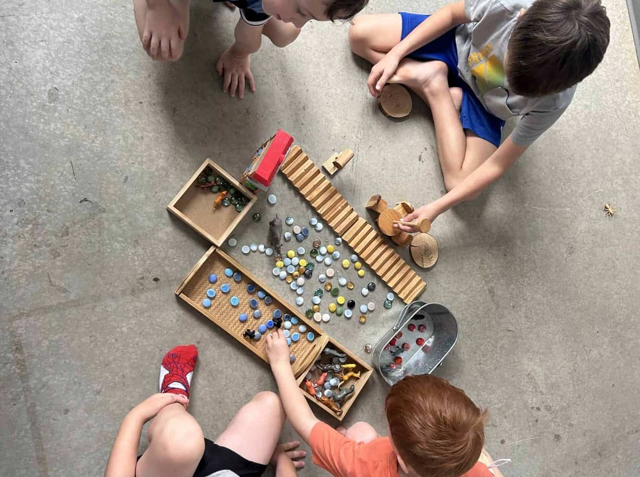 Four Selwyn students on the floor around wooden trays of glass marbles, a ridged wooden ramp and carved wooden vehicles, seen from above