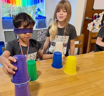 A student wearing a purple blindfold stacks a tower of coloured plastic cups while a classmate stands beside them giving directions.
