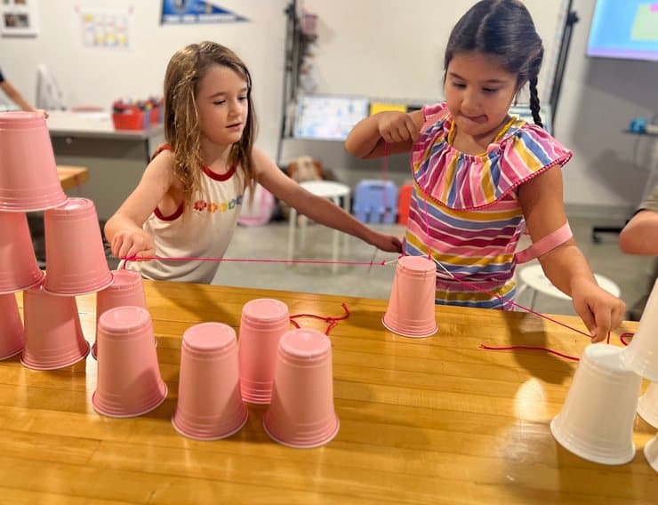Two students building a tall tower out of pink plastic cups on a classroom table.