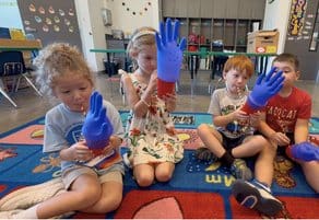 Students sitting on the classroom rug during a hands-on activity, two of them wearing bright blue gloves.