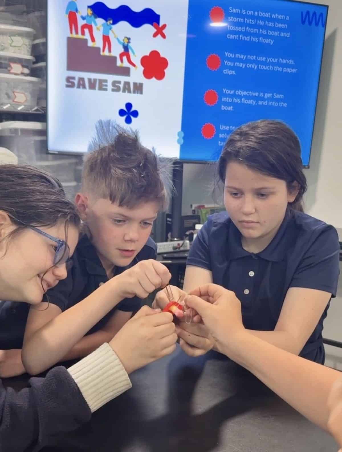 Three older Selwyn students leaning in over a table, working a paper clip and a small figure through a team challenge together, the rules of the challenge on the screen behind them.